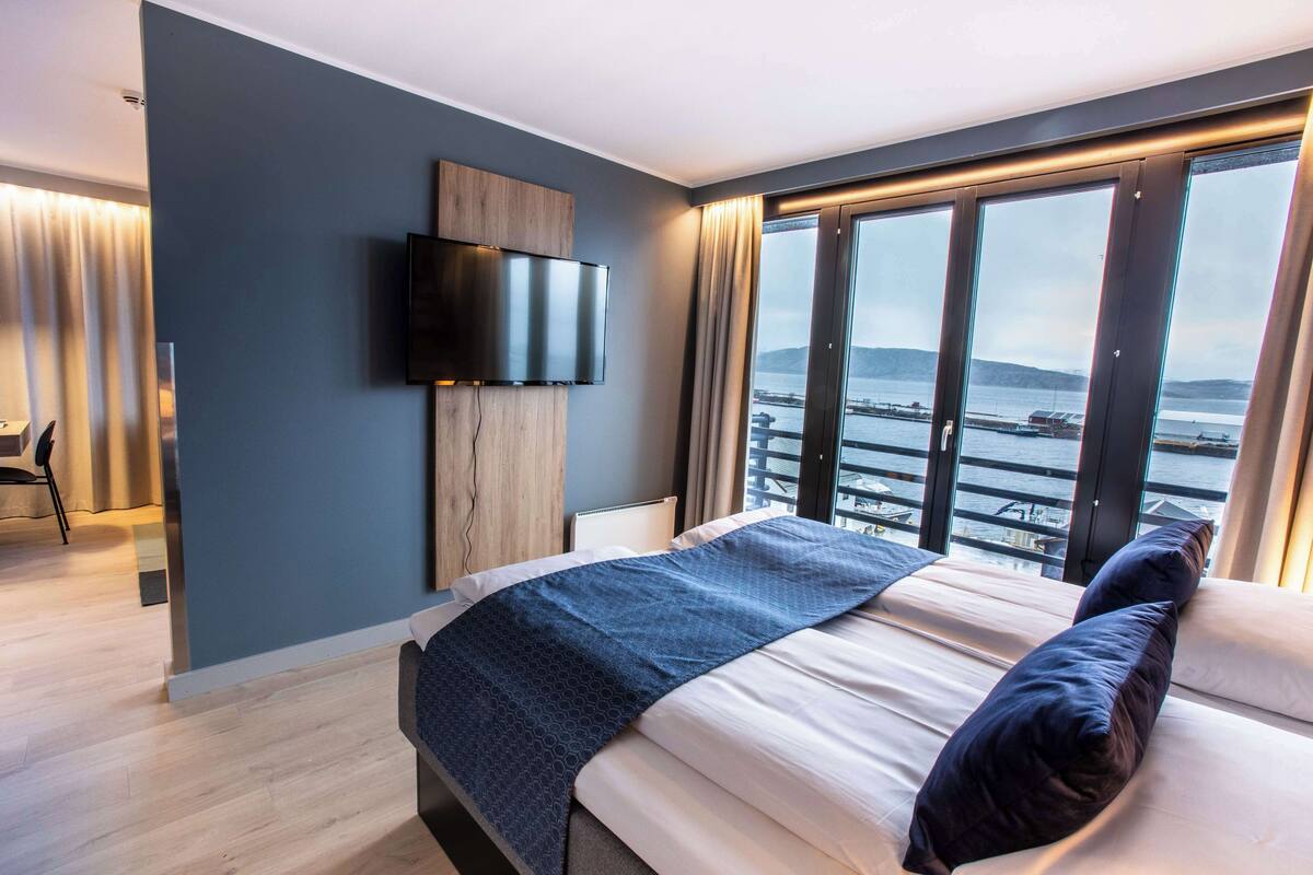 Une chambre d’hôtel moderne dotée d’un grand lit, d’un téléviseur à écran plat fixé au mur et offrant une vue sur un plan d’eau grâce à de grandes fenêtres.