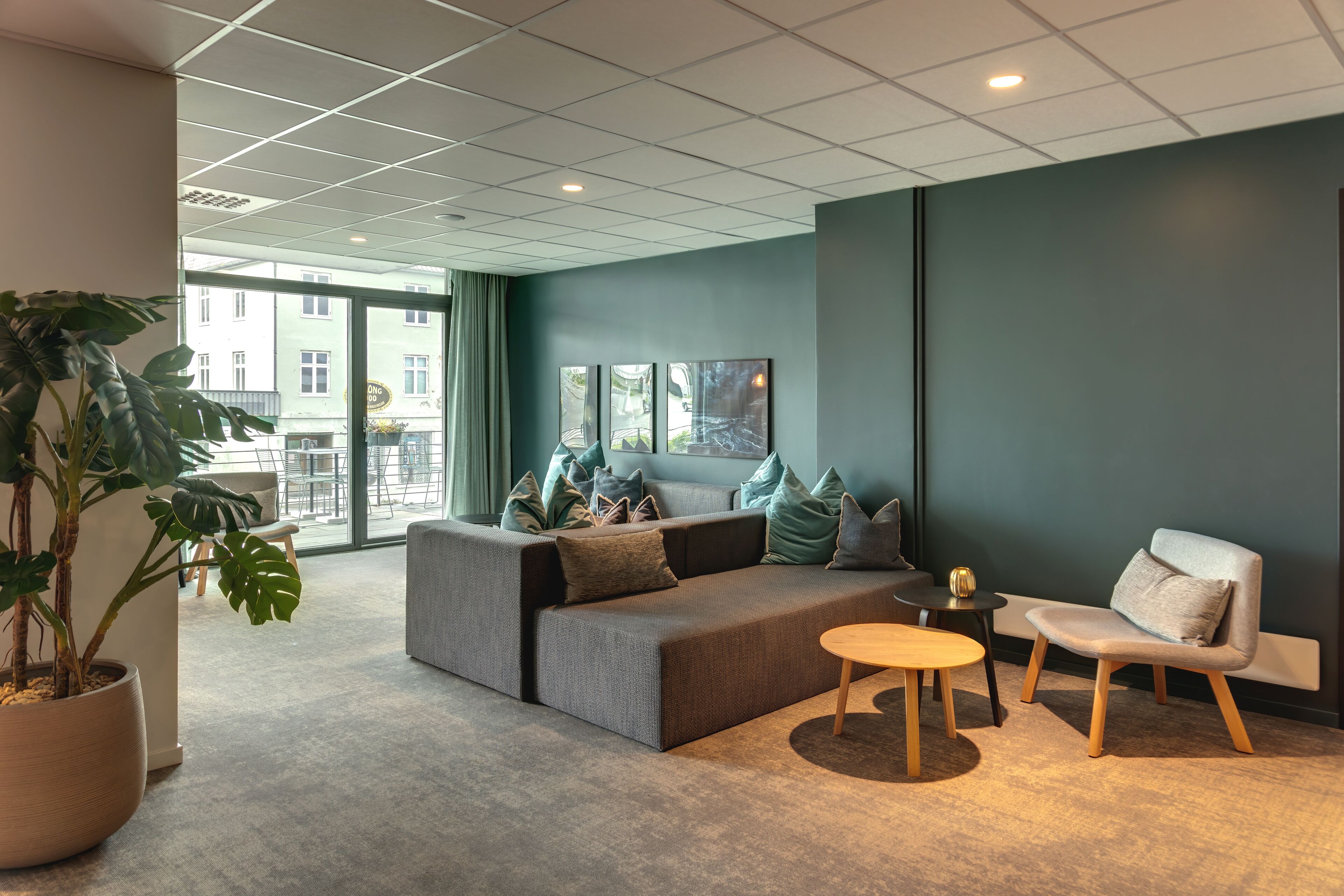 Un espace de détente moderne avec un canapé gris, une table ronde en bois et un banc. On y trouve une grande plante en pot et une œuvre d’art encadrée accrochée au mur.