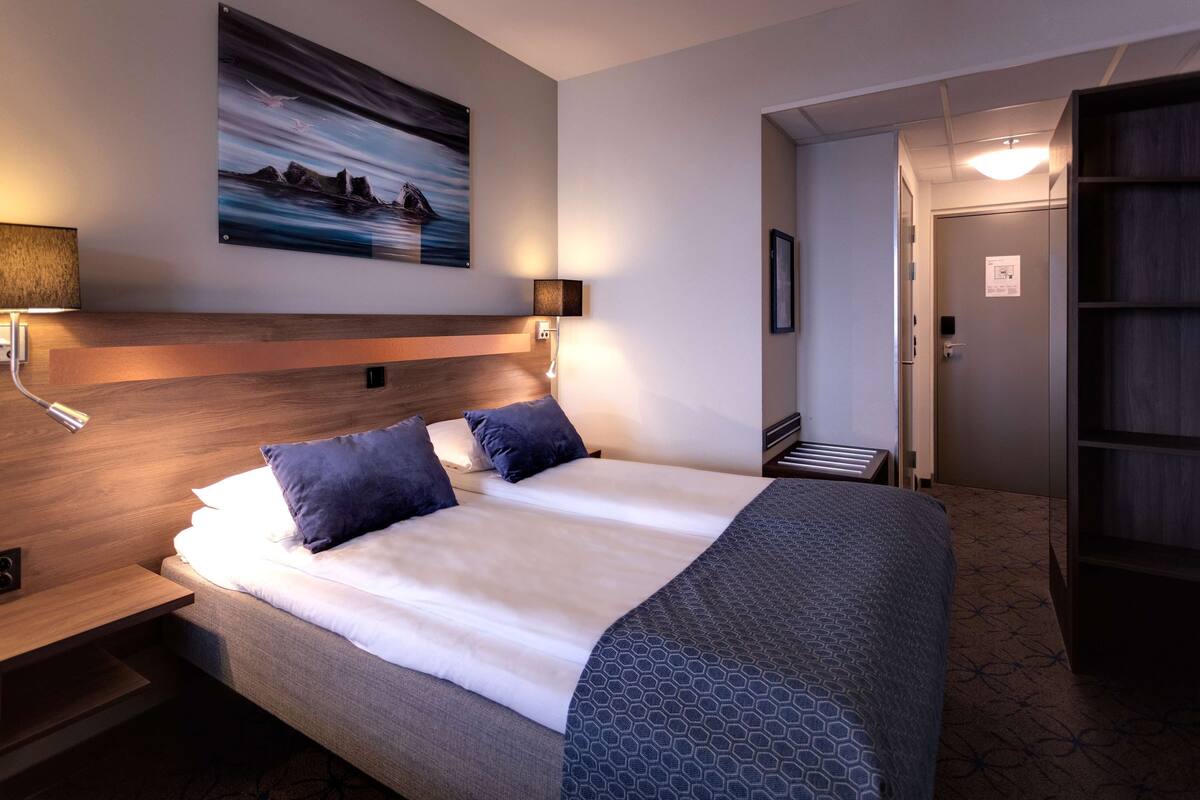 Une chambre d’hôtel avec un grand lit, une tête de lit, un tableau accroché au mur, une table de chevet avec une lampe et une porte donnant sur une autre pièce.