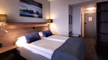 A hotel room with a large bed, a headboard, a wall-mounted painting, a bedside table with a lamp, and a door leading to another room.