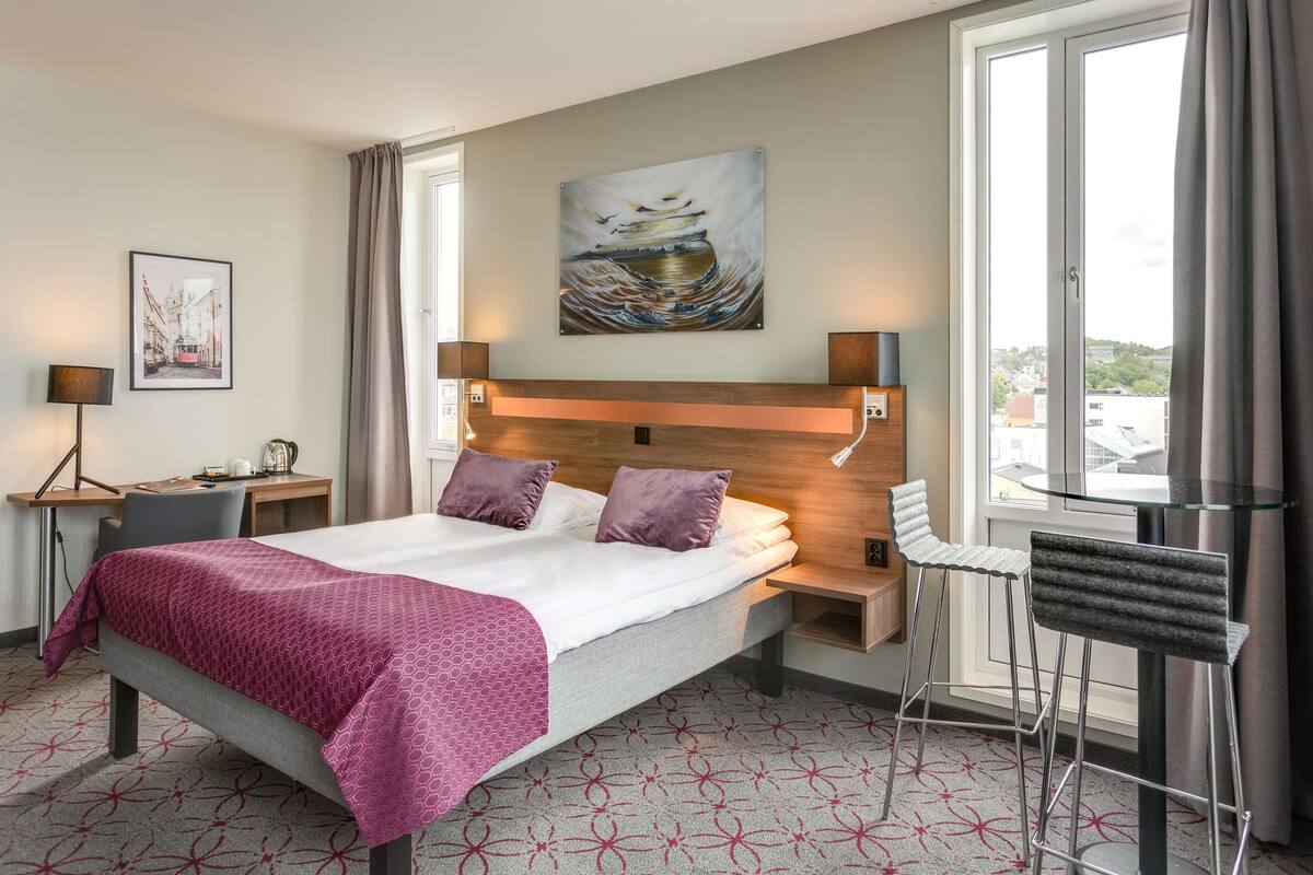 Une chambre d’hôtel avec un lit, un bureau, une chaise, une fenêtre avec des rideaux et des œuvres d’art accrochées au mur.