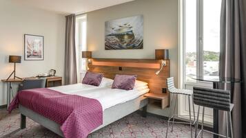 Ein Hotelzimmer mit Bett, Schreibtisch, Stuhl, einem Fenster mit Vorhängen und Bildern an der Wand.