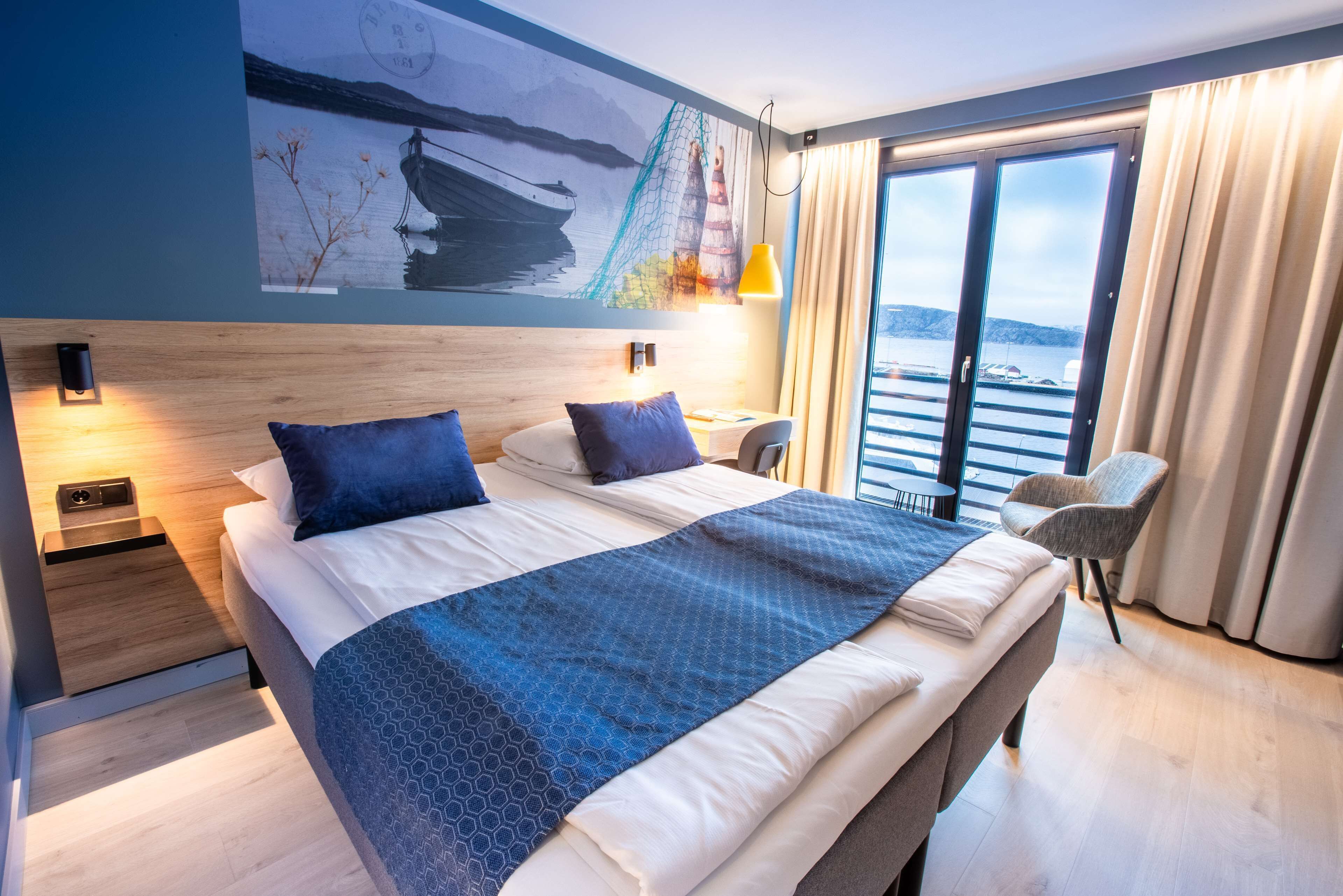 Una habitación de hotel moderna con una cama grande, un cuadro de un barco en la pared, vista al mar por la ventana y una silla junto a la ventana.