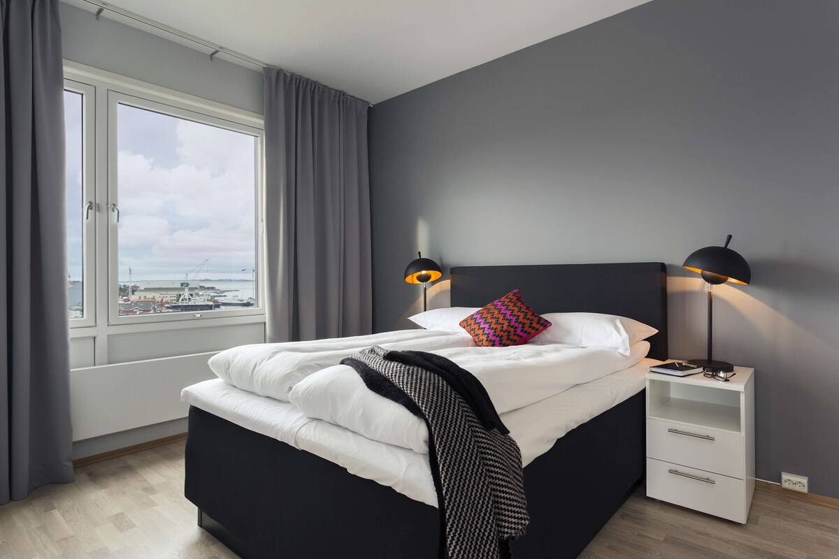 Une chambre à coucher moderne avec une grande fenêtre, un lit recouvert de draps blancs, une tête de lit noire, une table de chevet avec une lampe, et une vue sur le paysage urbain.
