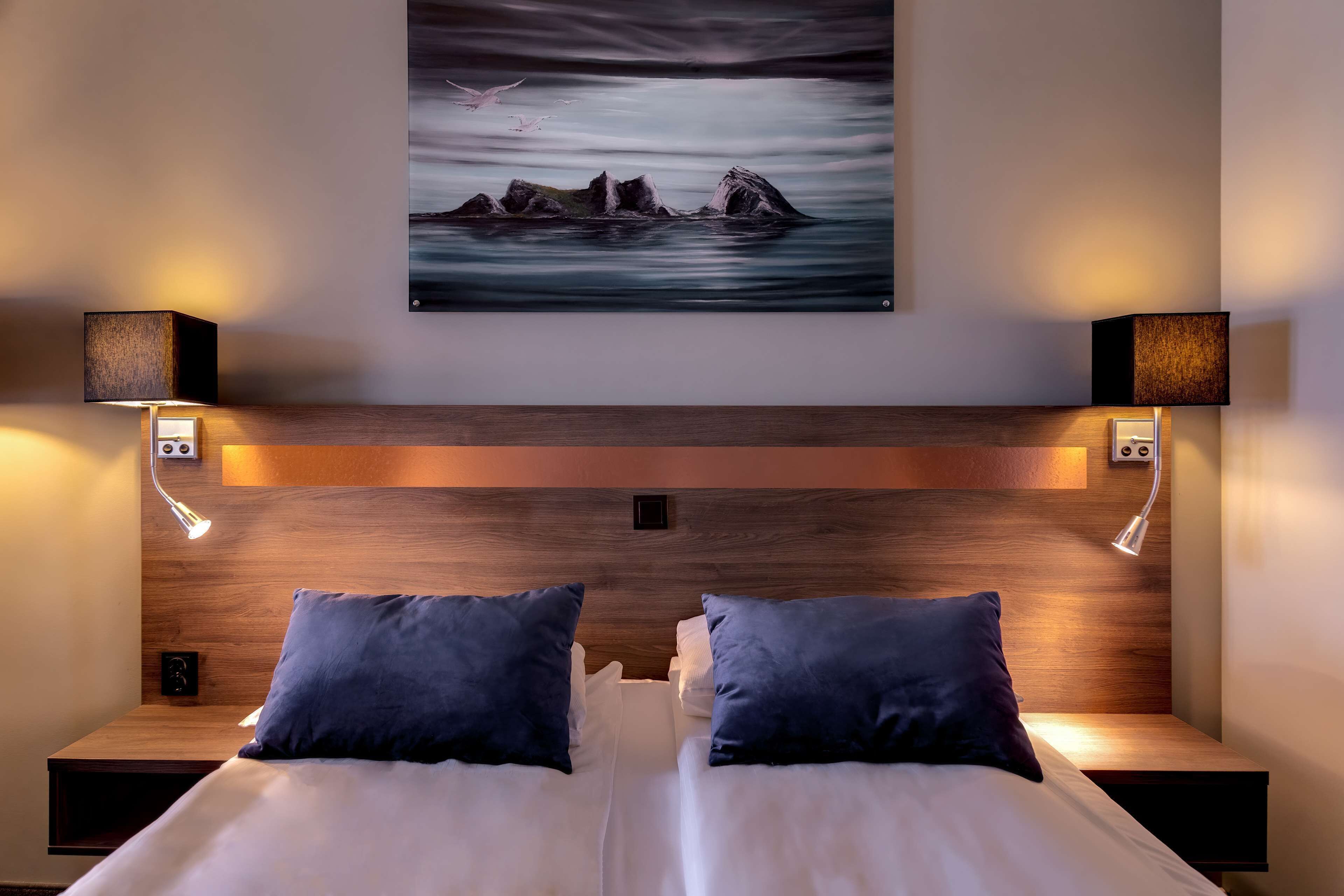 Habitación de hotel con cabecera de madera, dos mesitas de noche con lámparas, cama con ropa de cama blanca y almohadas oscuras, y una pintura de paisaje marino sobre la cabecera.