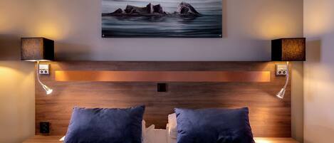 Habitación de hotel con cabecera de madera, dos mesitas de noche con lámparas, cama con ropa de cama blanca y almohadas oscuras, y una pintura de paisaje marino sobre la cabecera.