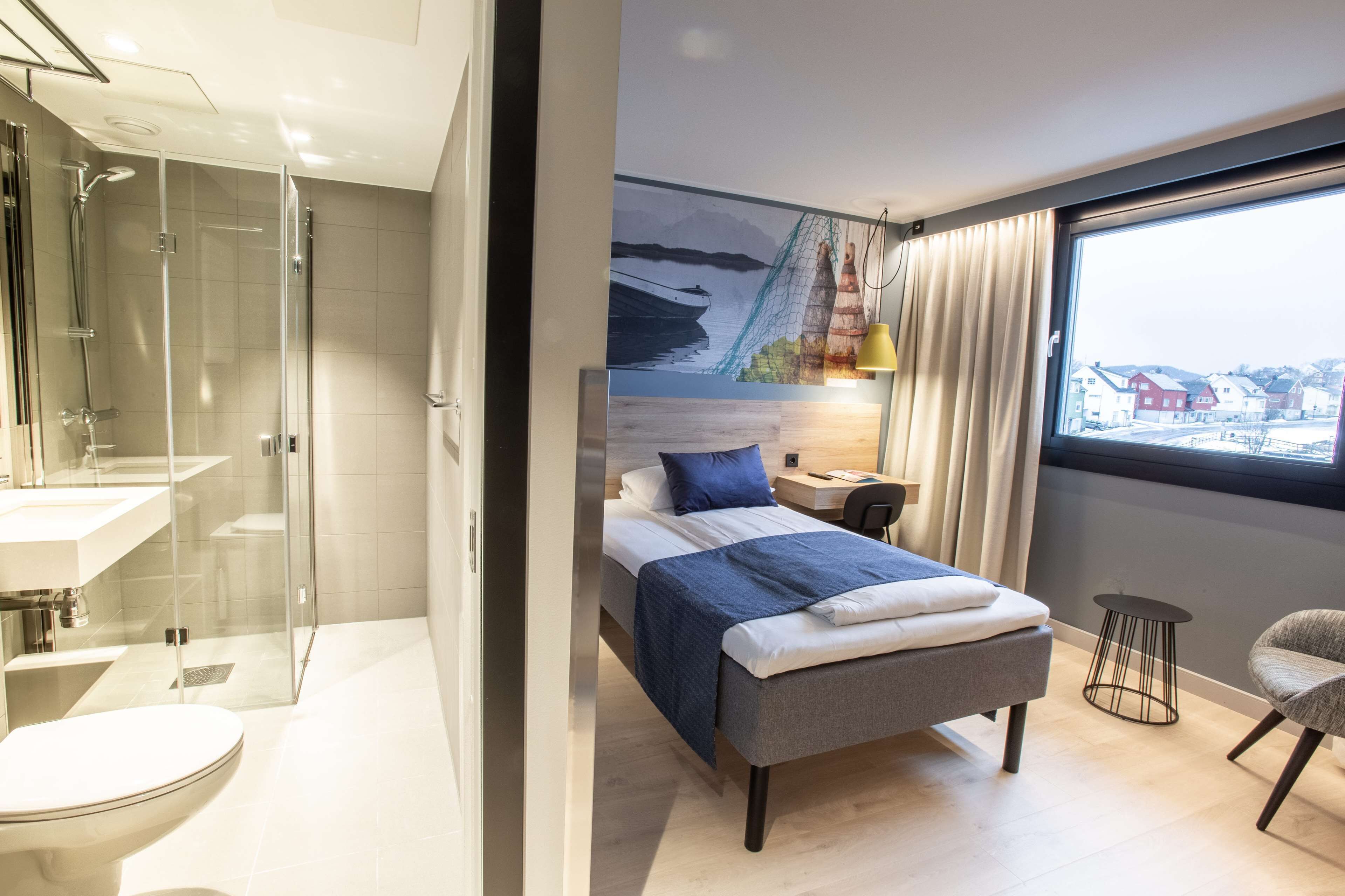 Une chambre d’hôtel moderne dotée d’une douche à parois de verre, d’un lit avec une tête de lit bleue, d’une table de chevet avec une lampe et d’une vue sur un paysage enneigé par une grande fenêtre.