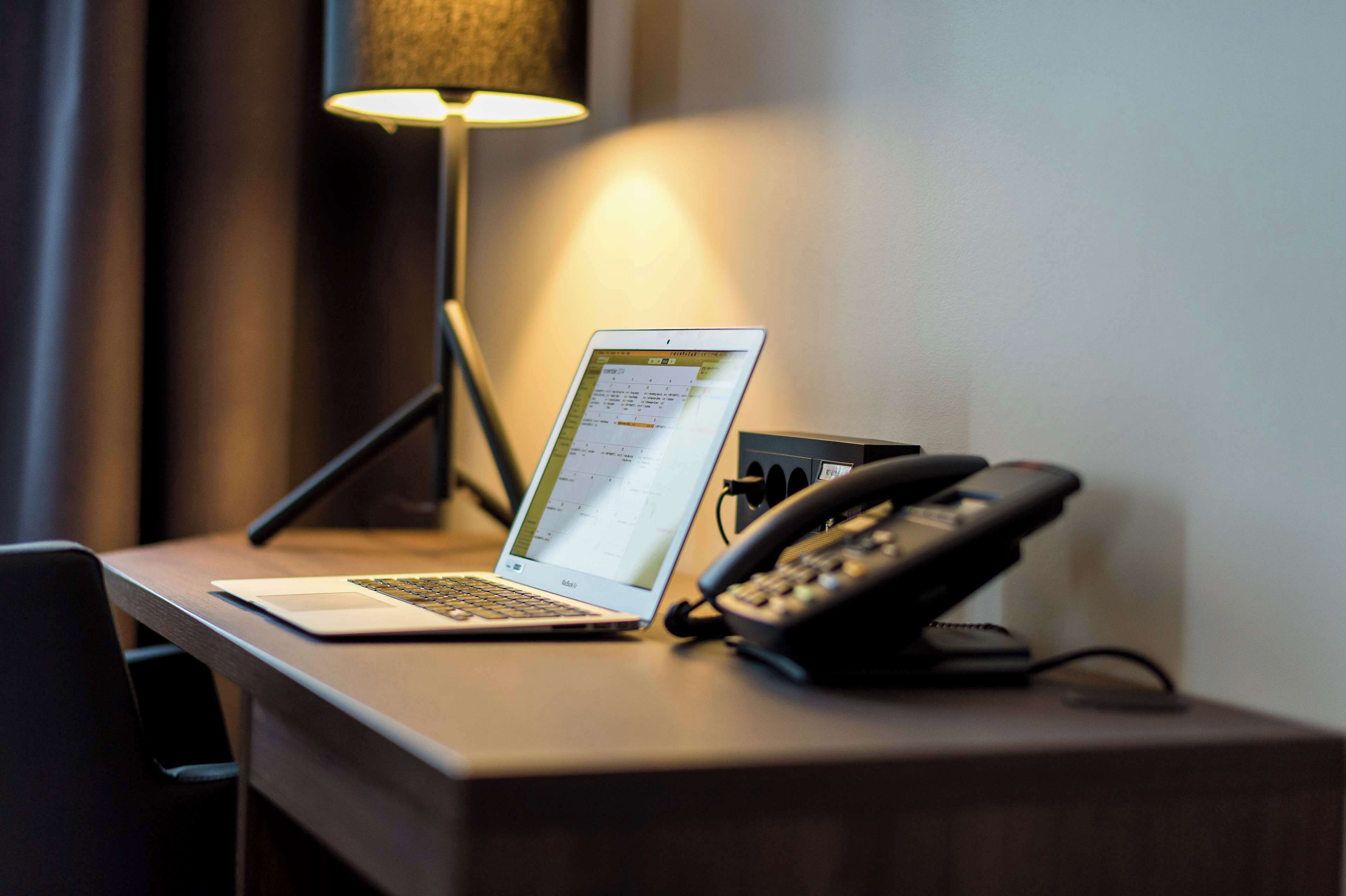 A laptop on a desk with a telephone and a lamp in the background.