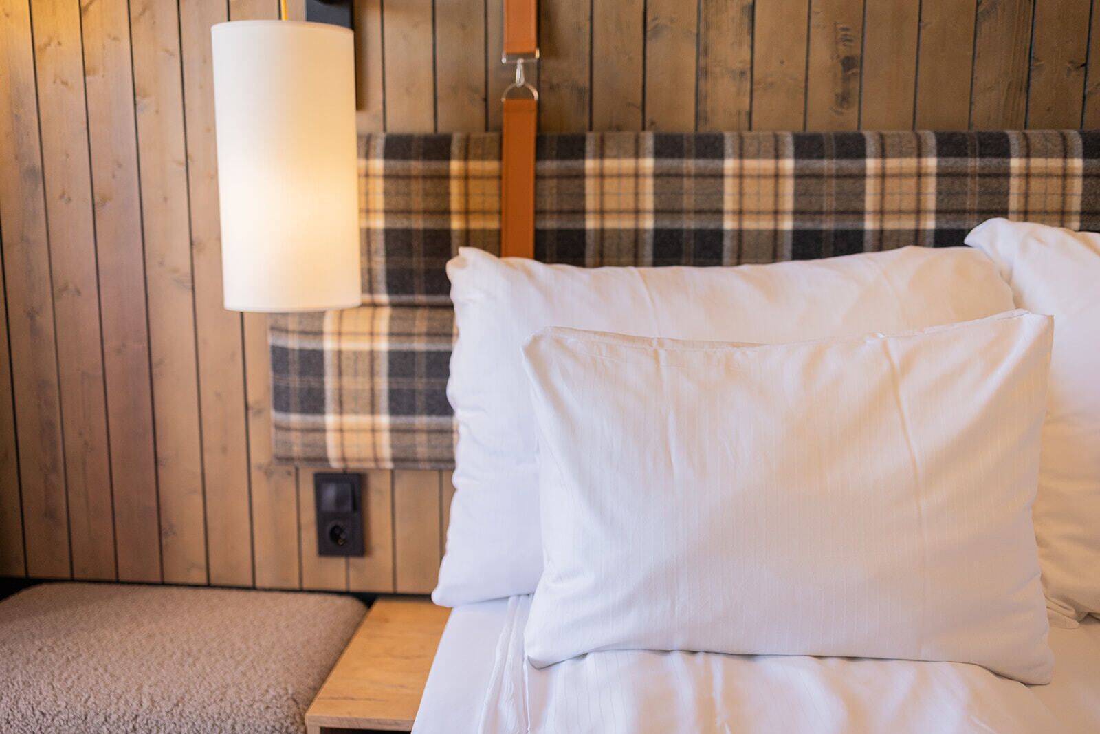 Une chambre avec un mur en bois, une tête de lit à carreaux, un lit blanc avec des oreillers, une table de chevet et une lampe.