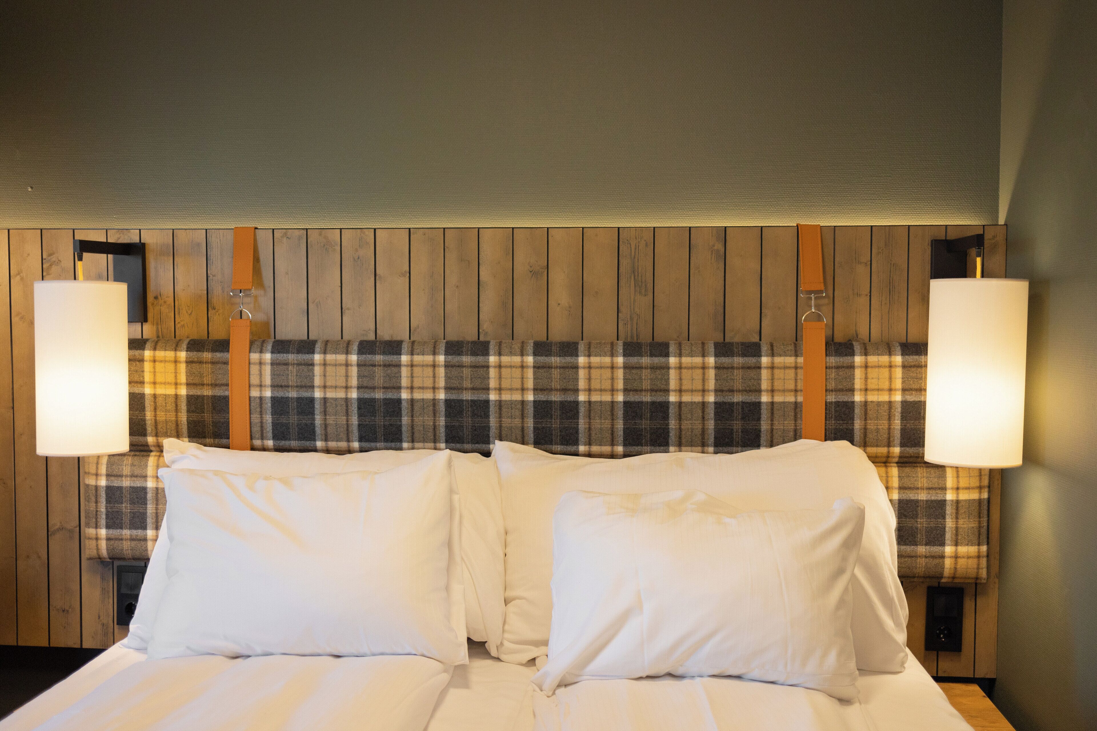 A hotel room with a wooden headboard, plaid headboard cover, and two bedside lamps.