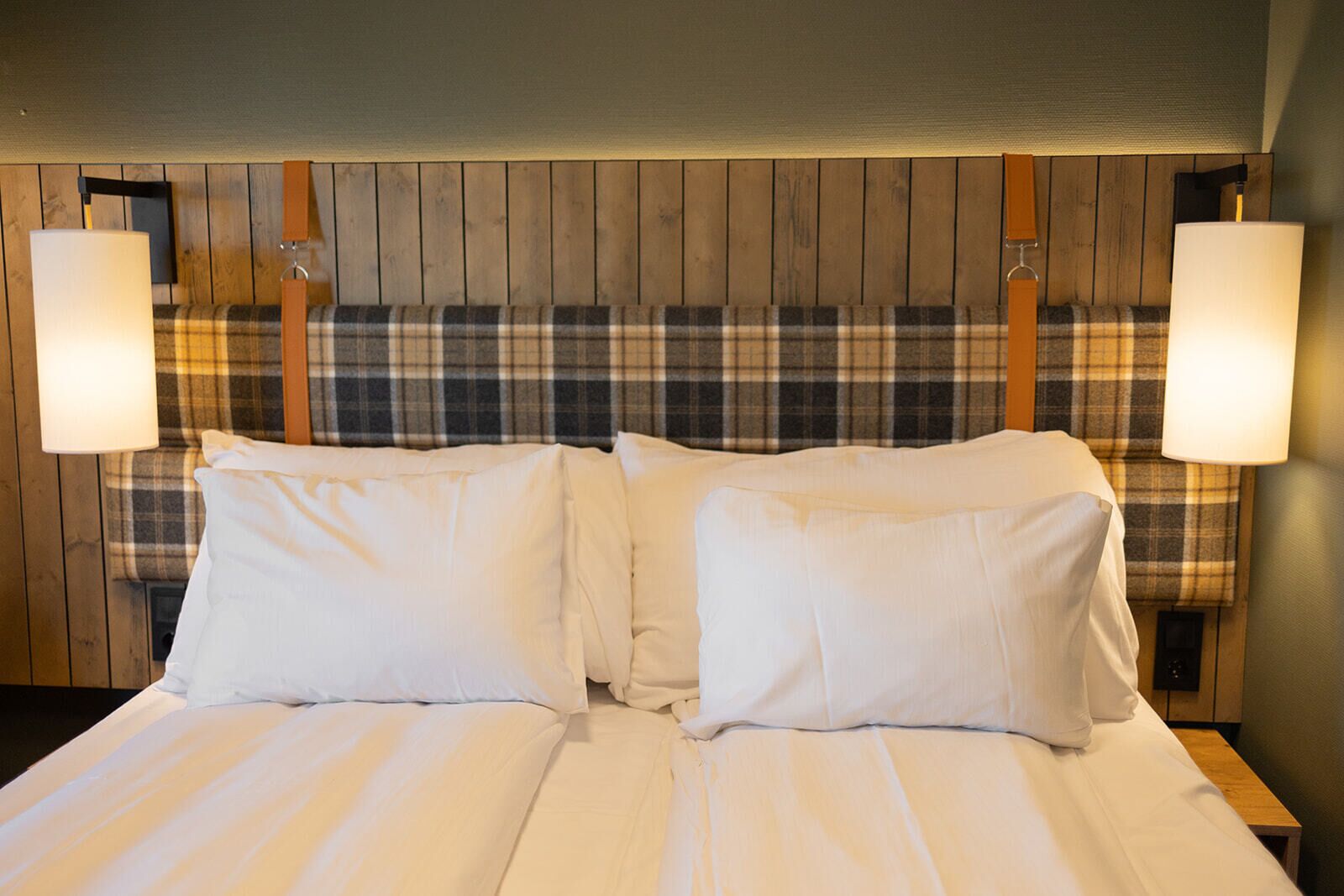 Una cama doble con sábanas blancas, flanqueada por dos lámparas de pared, en una habitación con paneles de madera y un cabecero estampado de cuadros.