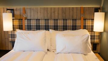 Una cama doble con sábanas blancas, flanqueada por dos lámparas de pared, en una habitación con paneles de madera y un cabecero estampado de cuadros.