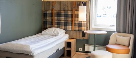 Habitación de hotel compacta con una cama individual, una mesita de noche pequeña, una silla y una mesa redonda.