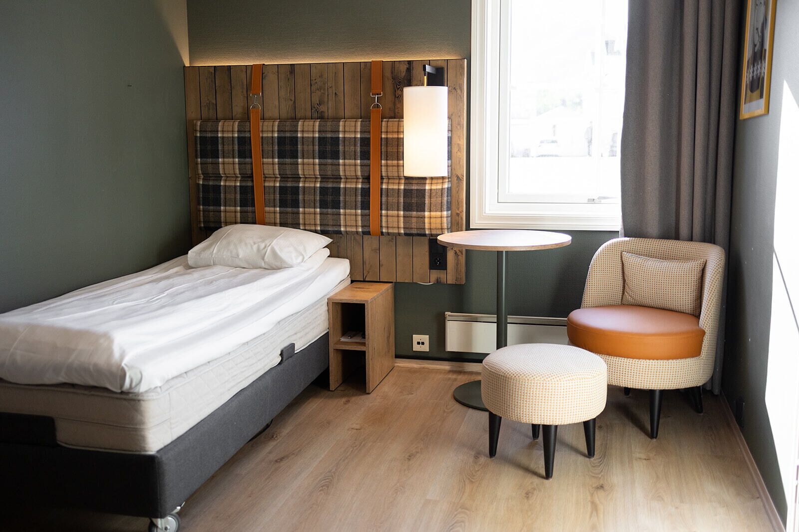 A compact hotel room with a bed, a small round table, a chair, and a window with curtains.