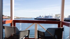 Vista de uma varanda com duas cadeiras de vime, com vista para um corpo d'água calmo e um grande navio atracado em um cais.