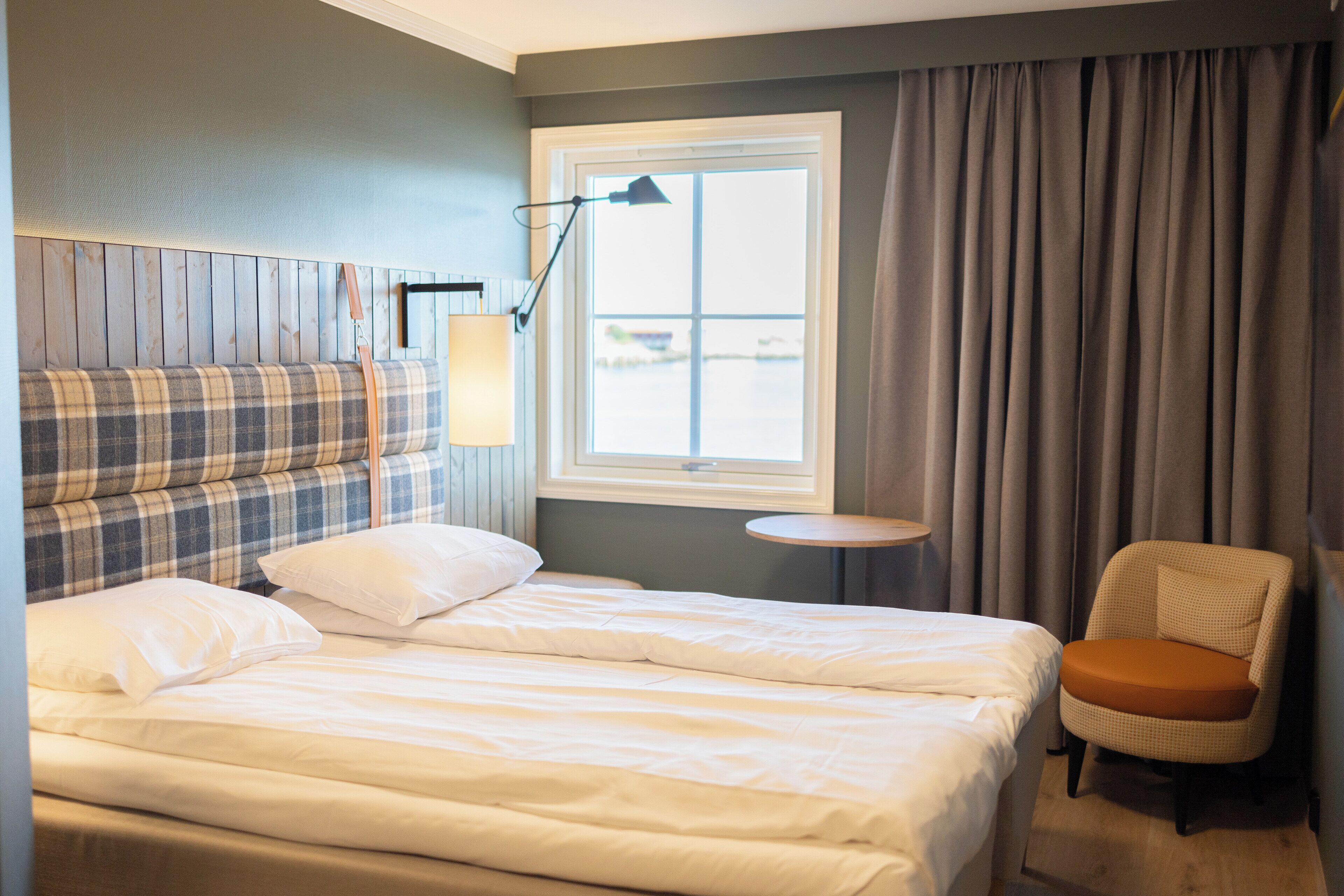 Habitación de hotel con una cama doble, una mesita pequeña y redonda, una silla con cojín naranja, una lámpara y una ventana con cortinas.