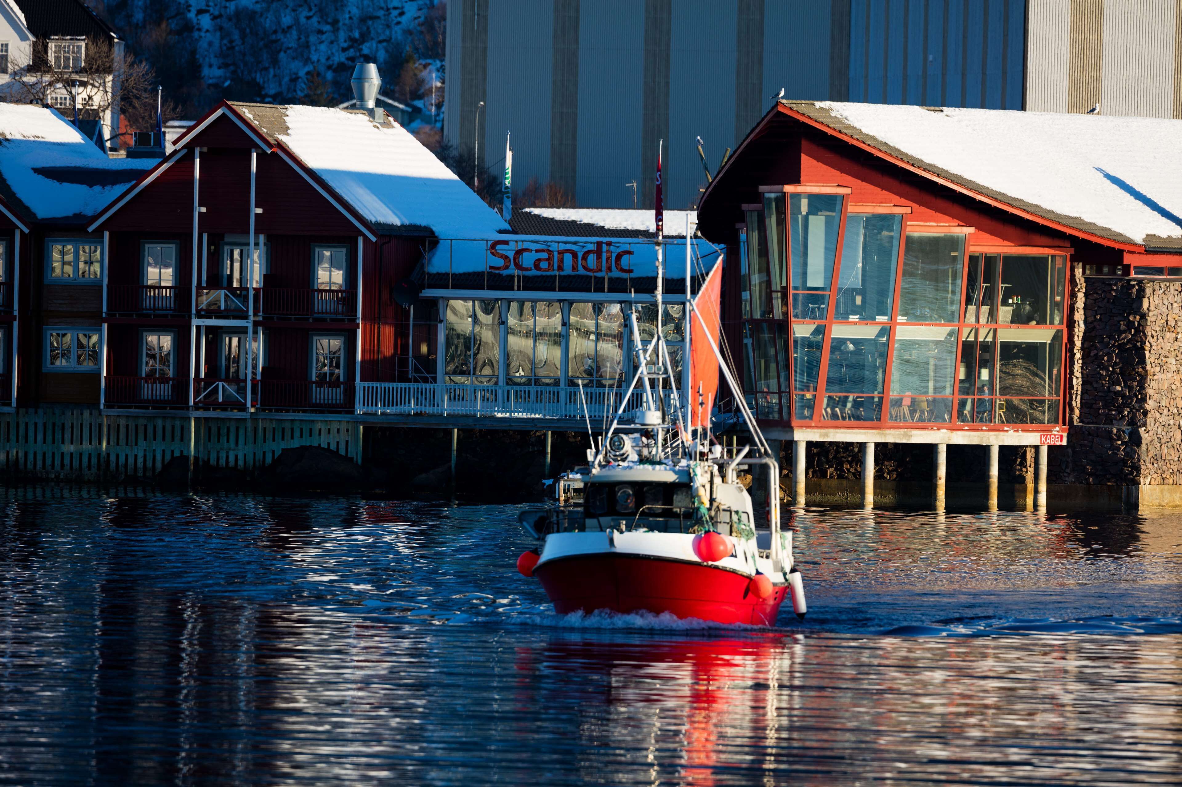 Um barco vermelho e branco atracado em um cais, com um edifício com a inscrição 'Scandic' ao fundo.