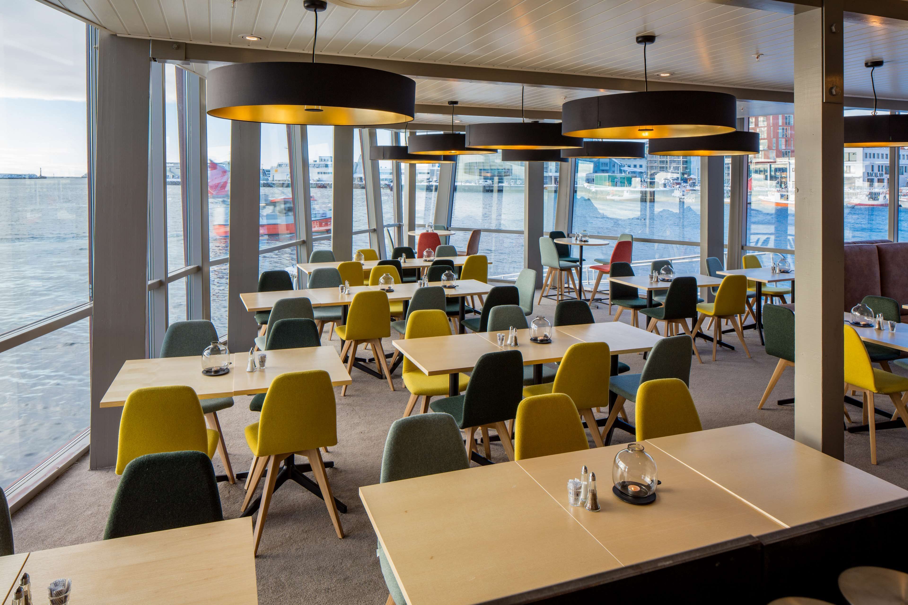 A dining area with tables and chairs, large windows with a view of the water and buildings, and pendant lights.