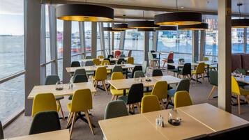 A dining area with tables and chairs, large windows with a view of the water and buildings, and pendant lights.