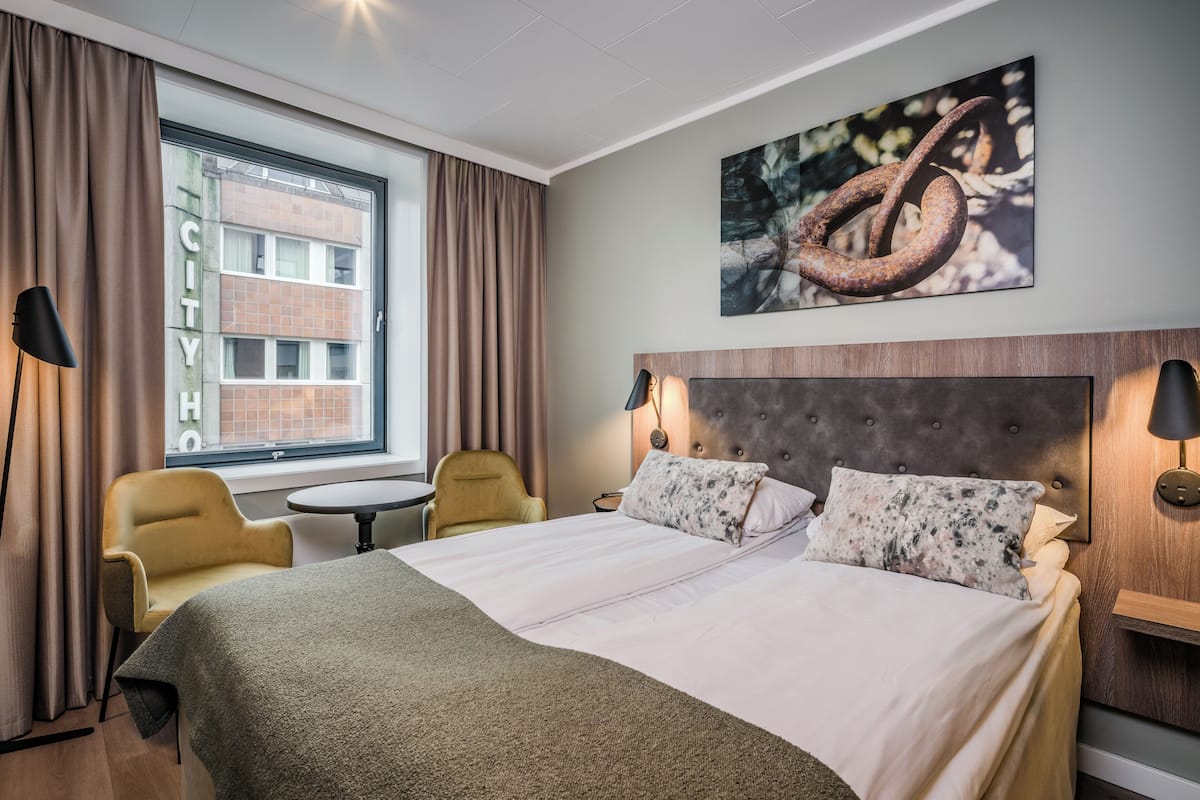 Ett hotellrum med en stor säng, två sänglampor, ett runt bord, en stol och ett fönster med gardiner.