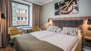 A hotel room with a large bed, two bedside lamps, a round table, a chair, and a window with curtains.