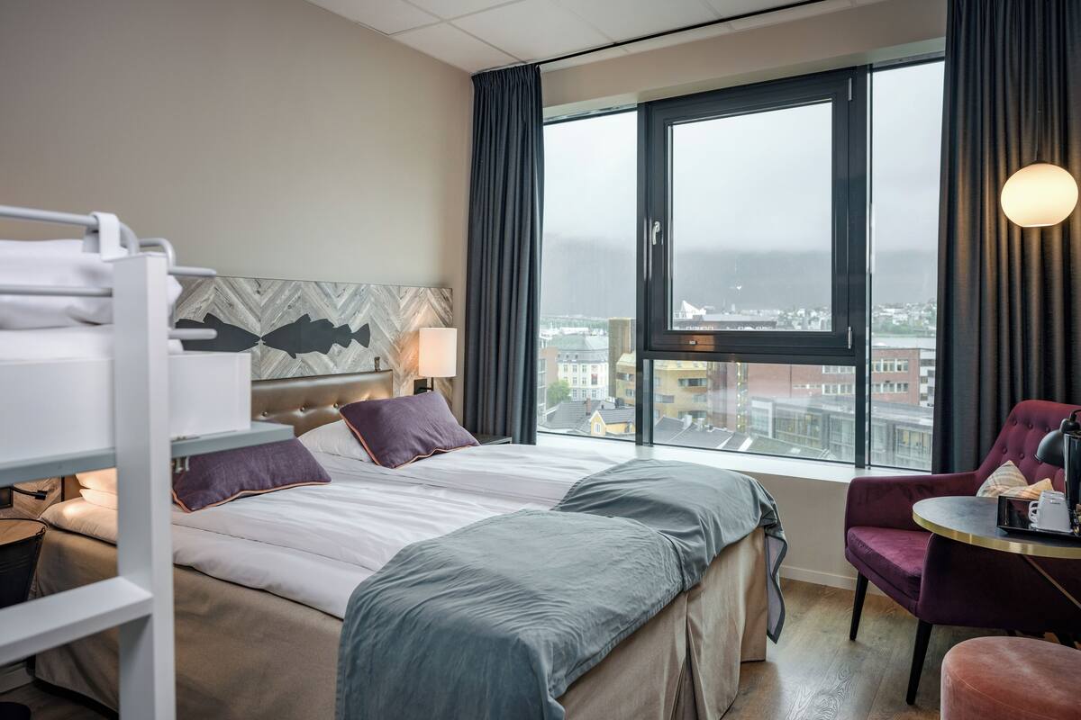 Ett hotellrum med en säng, ett skrivbord, en stol, ett fönster med utsikt över staden och en våningssäng.