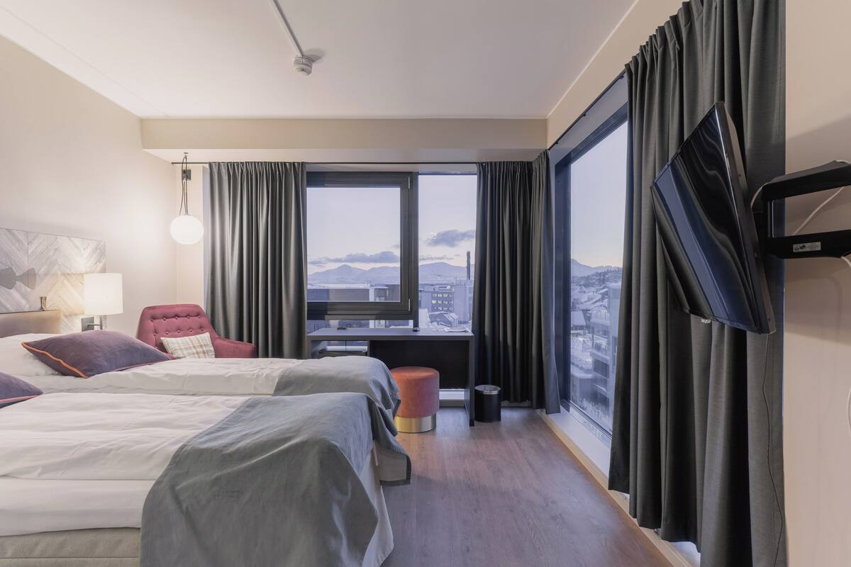 Ett hotellrum med två sängar, en röd stol, ett skrivbord med en tv och ett stort fönster med gardiner.