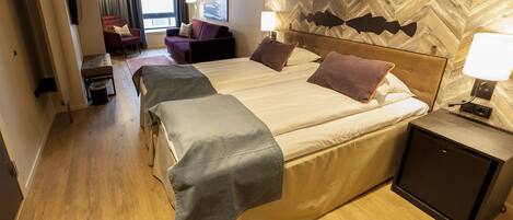 Ein Hotelzimmer mit einem großen Bett, zwei Nachttischen, einem kleinen Schreibtisch, einem Sessel, einem Fernseher und einem Fenster mit Vorhängen.