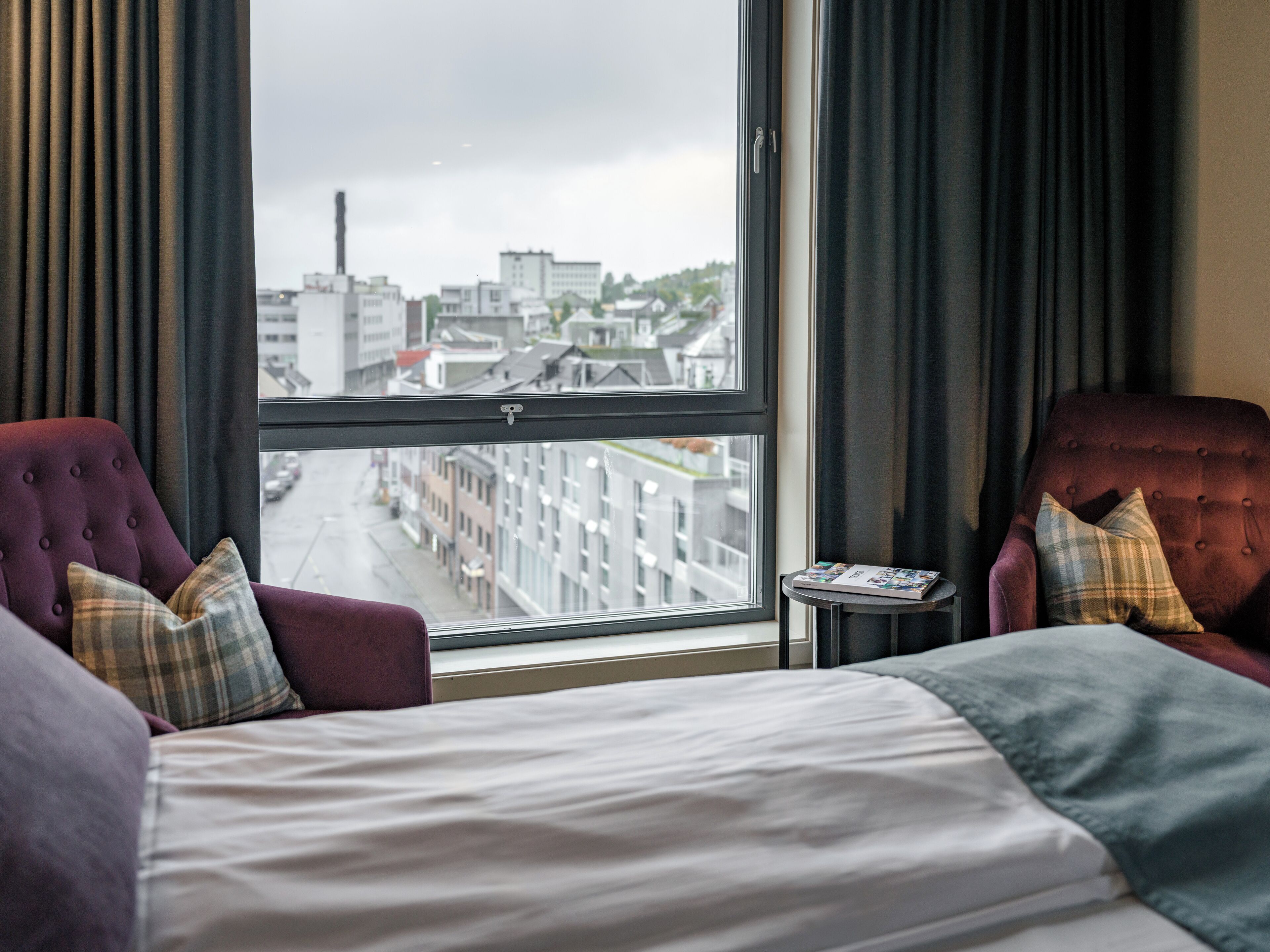 A hotel room with a large window, two armchairs, a small table, and a bed.