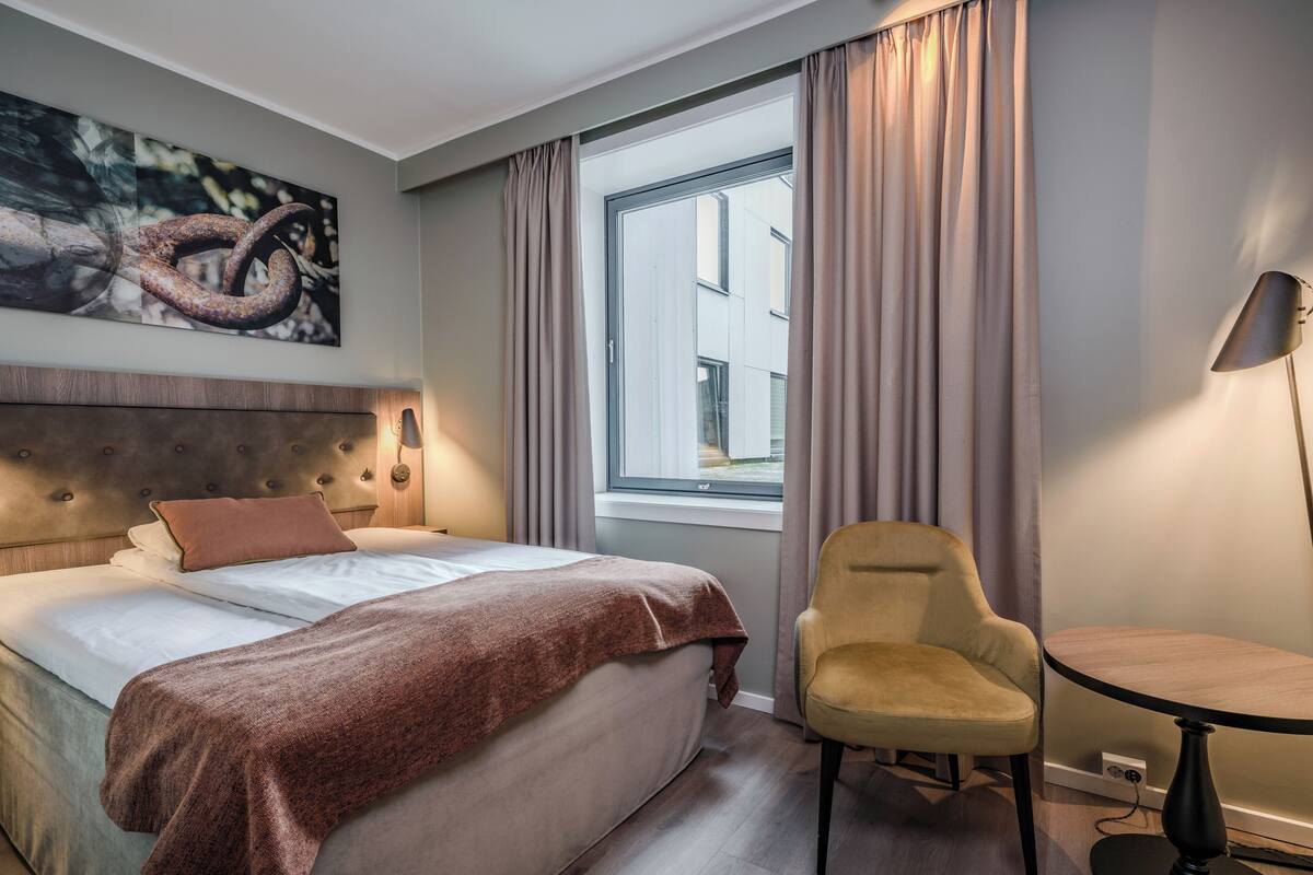 Ett hotellrum med en säng, en stol, ett skrivbord, ett fönster med gardiner och en tavla på väggen.