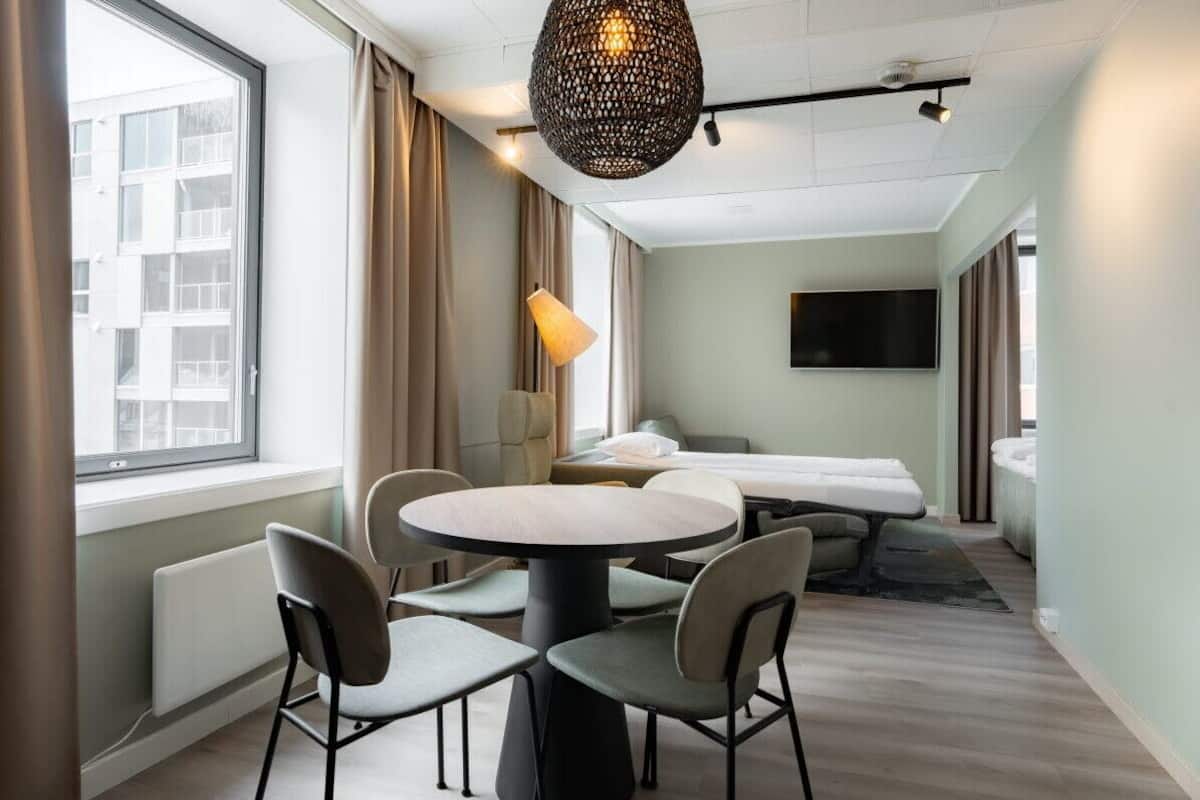 Ett hotellrum med ett runt bord, fyra stolar, en säng, en tv och ett stort fönster med gardiner.