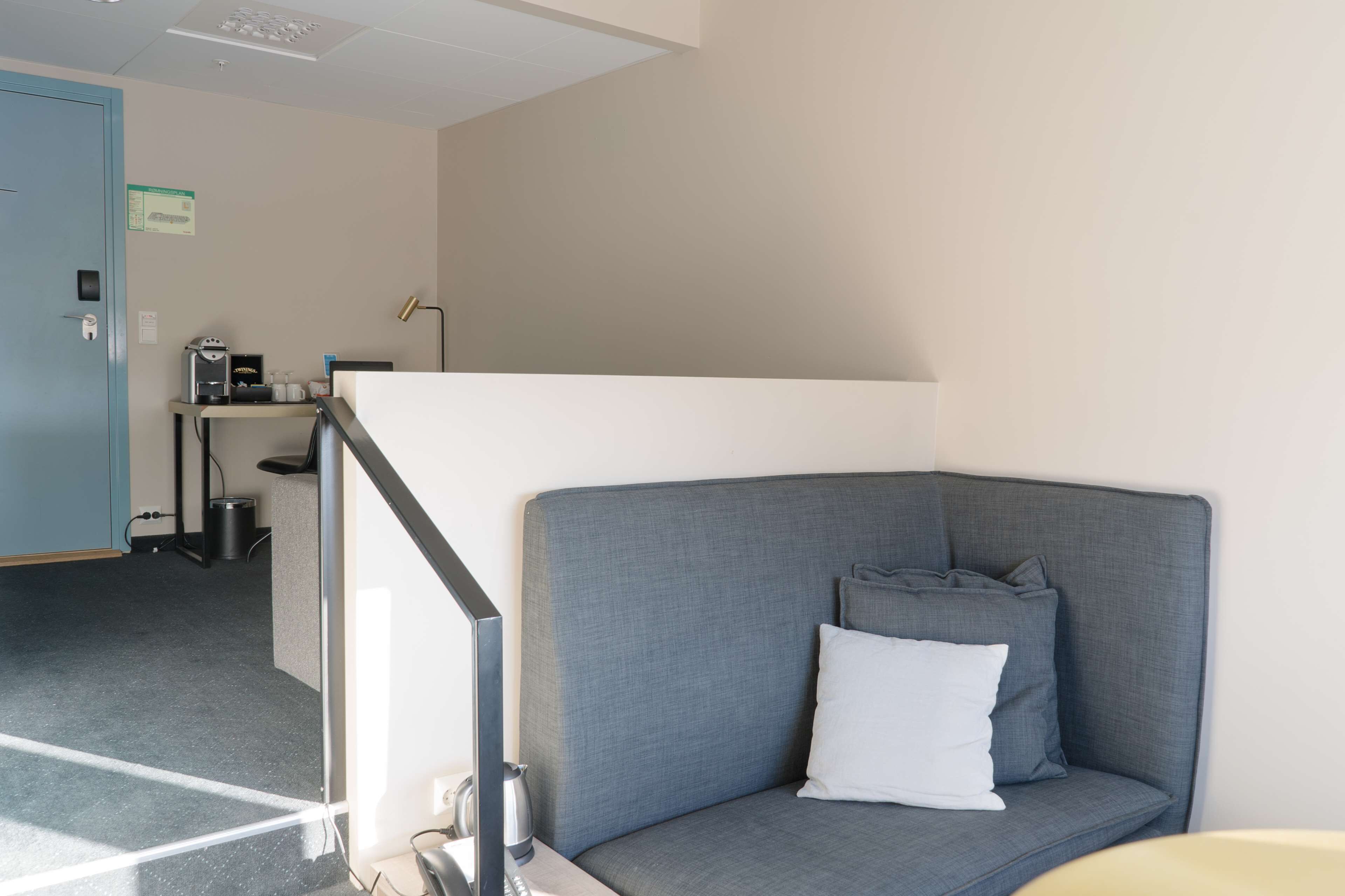 A modern hotel room with a grey sofa, a white pillow, a small table, and a desk with a lamp.