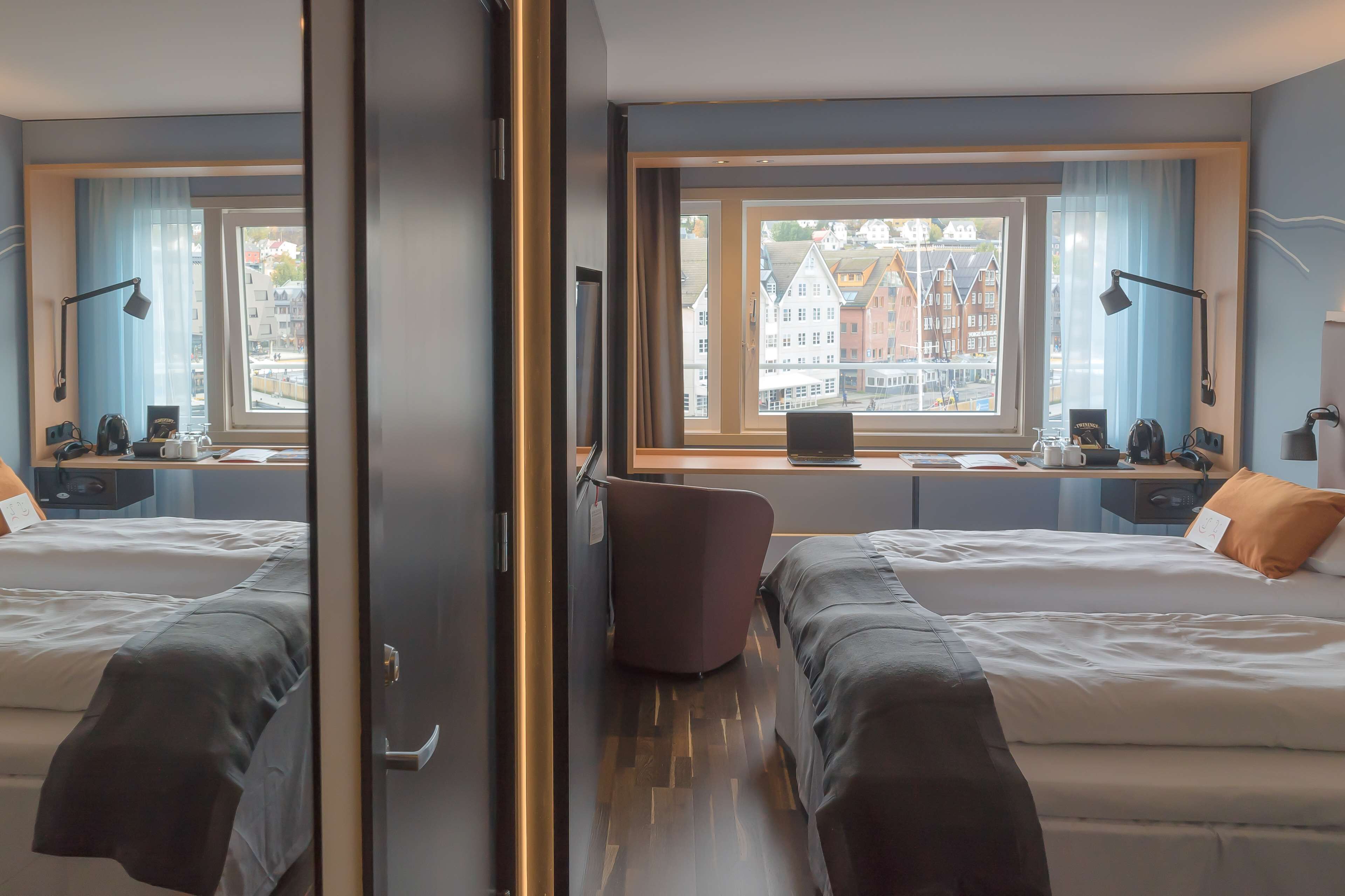 Hotellihuone, jossa on kaksi sänkyä, työpöytä, tuoli ja suuri ikkuna, josta on näkymä rakennuksiin.