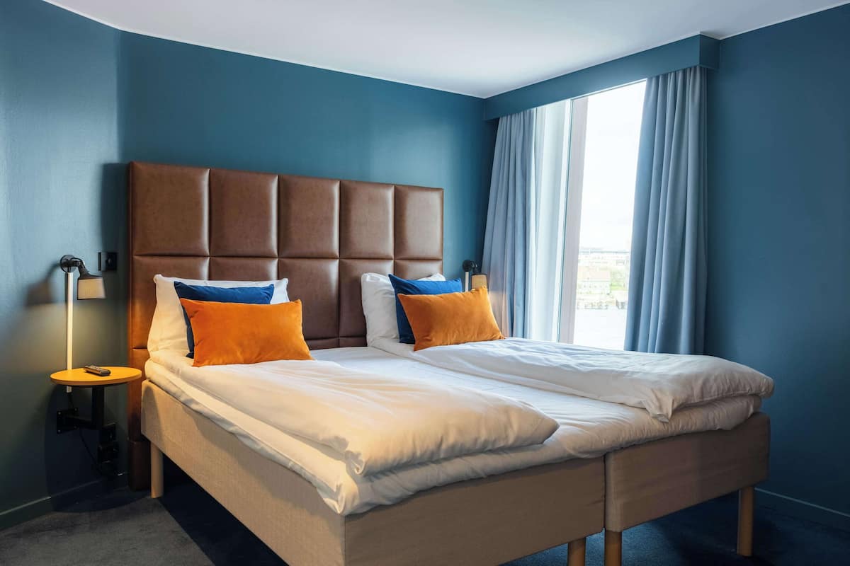 A double bed with a padded headboard, two pillows, a bedside table with a lamp, and a large window with curtains.