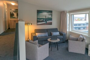 A modern living room with a grey sofa, two armchairs, a coffee table, and a laptop. A painting of a woman by the sea hangs on the wall.