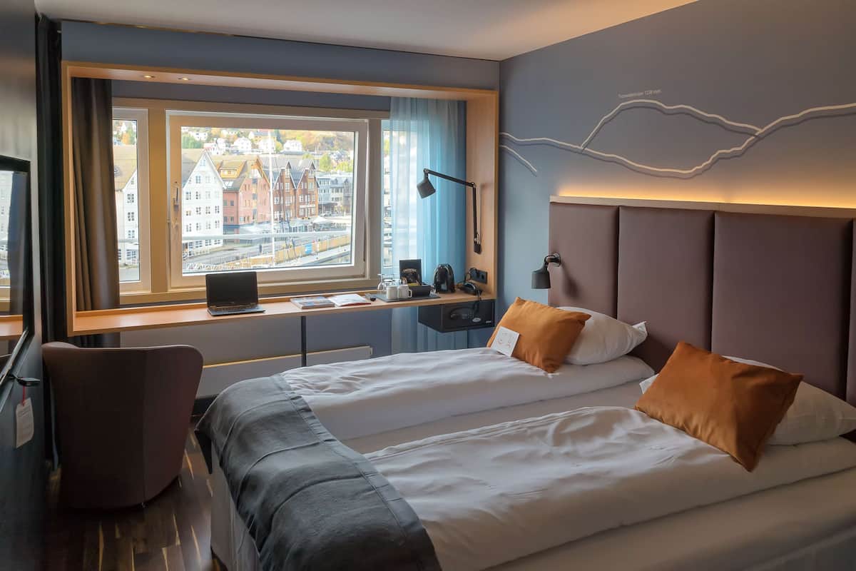 A hotel room with a large bed, a desk with a laptop, and a view of buildings outside the window.