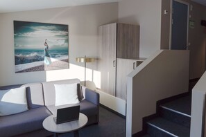 A modern living room with a sofa, a laptop, a painting of a woman by the sea, a lamp, and a staircase.