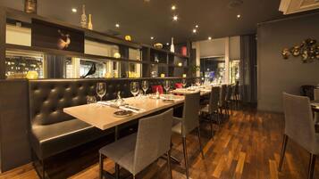Un restaurante con una larga mesa dispuesta para comer, un banco acolchado negro y vistas de la ciudad a través de amplios ventanales.