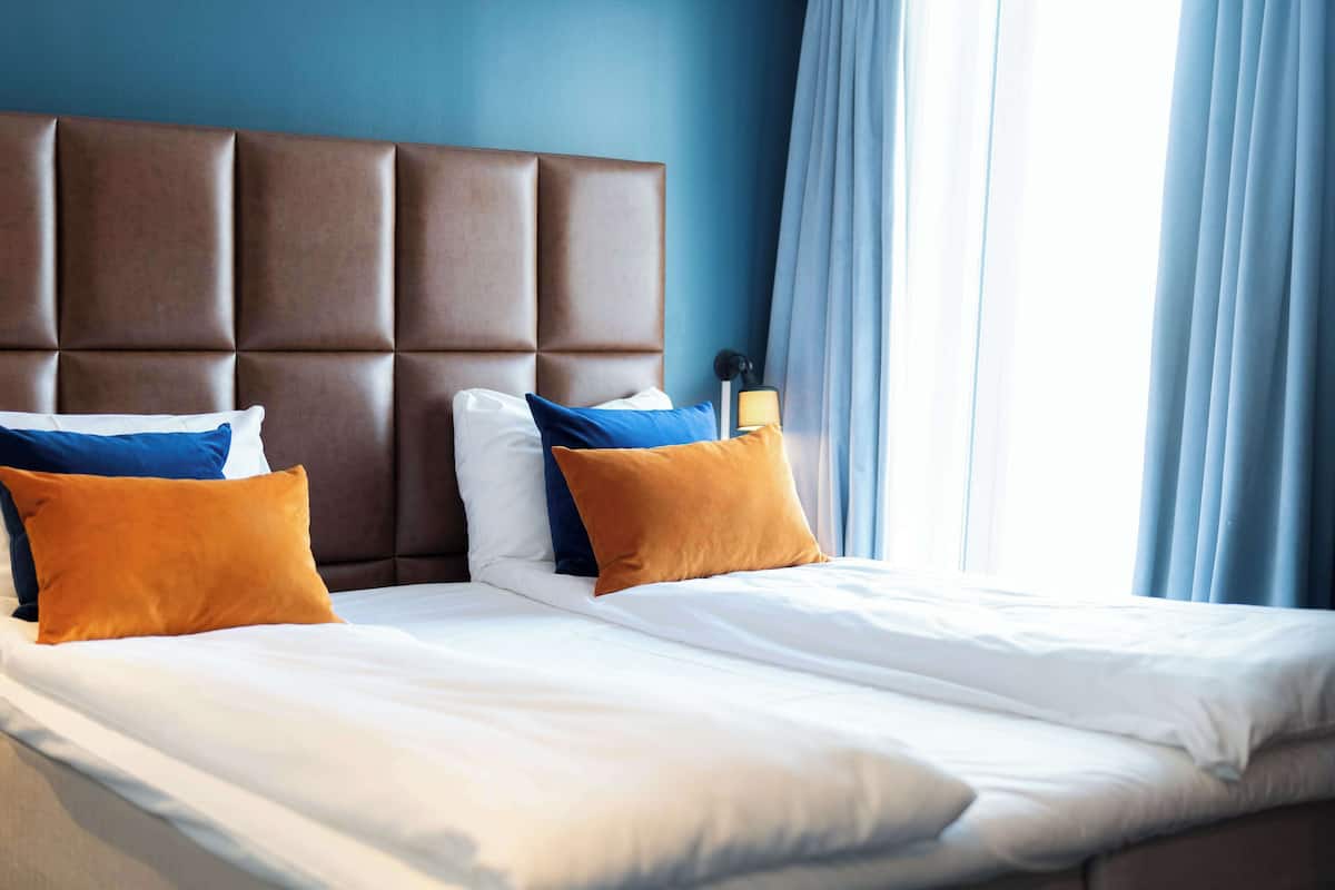 A hotel room with a large padded headboard, two beds, and a window with blue curtains.