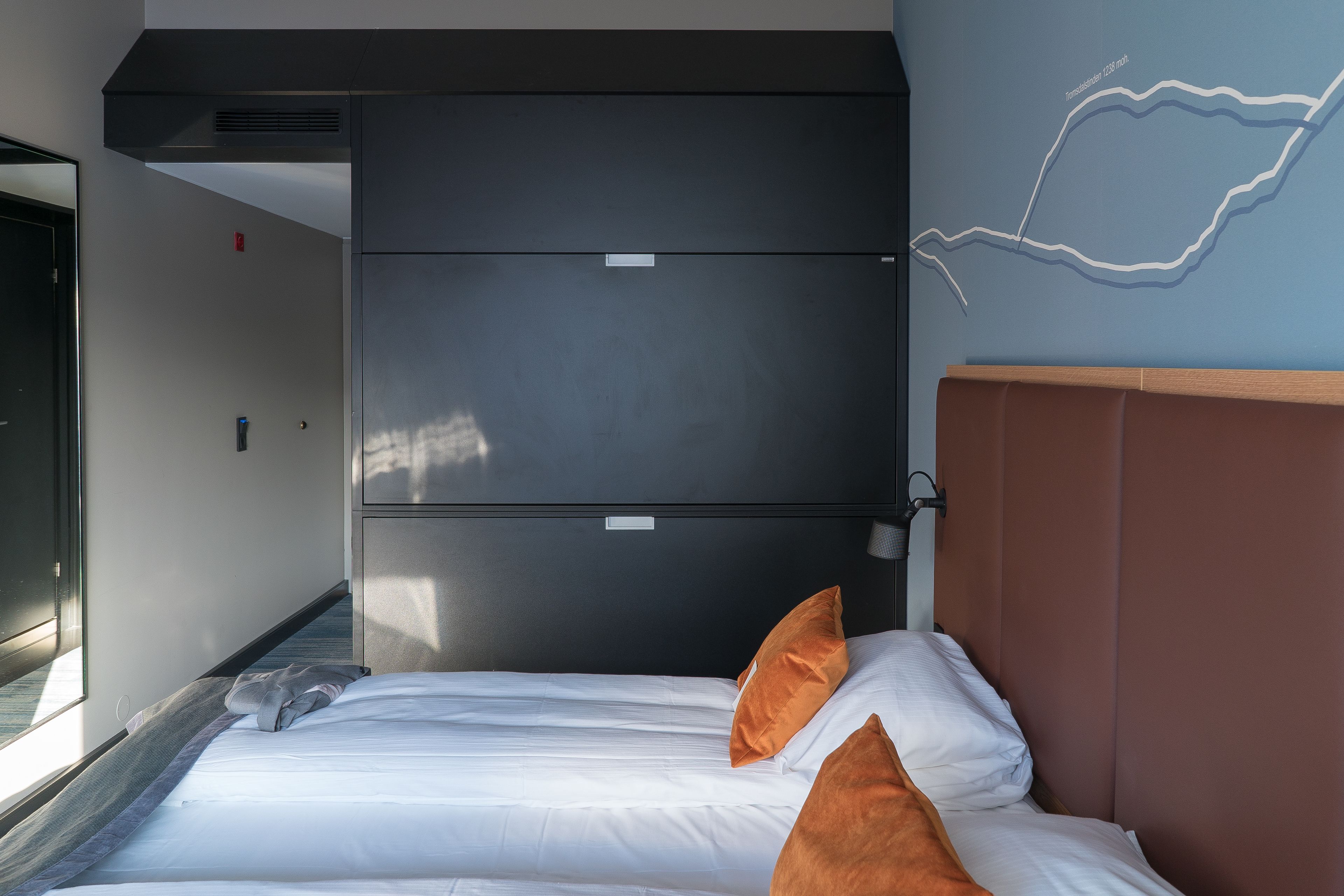 A hotel room with a bed, a headboard, a wall-mounted heater, and a window.
