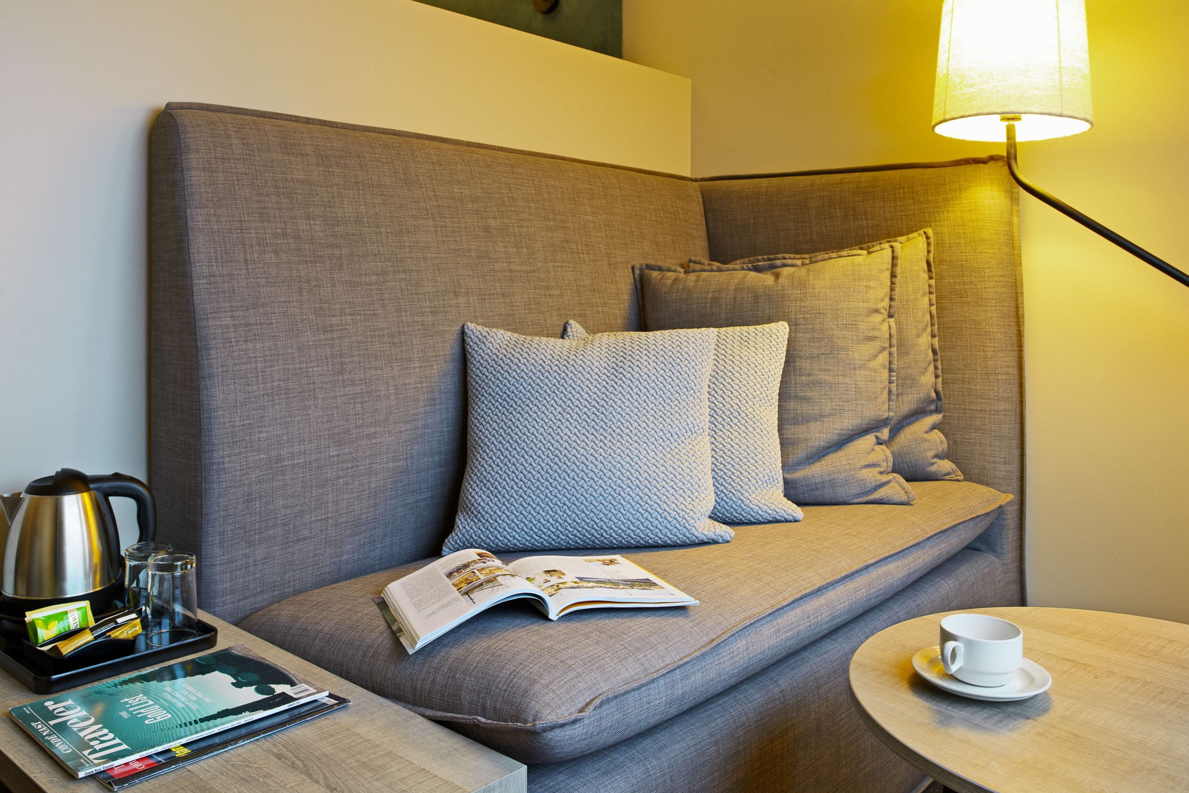 A modern hotel room with a sofa, a magazine, a tray with a kettle, glasses, and a cup on a side table.