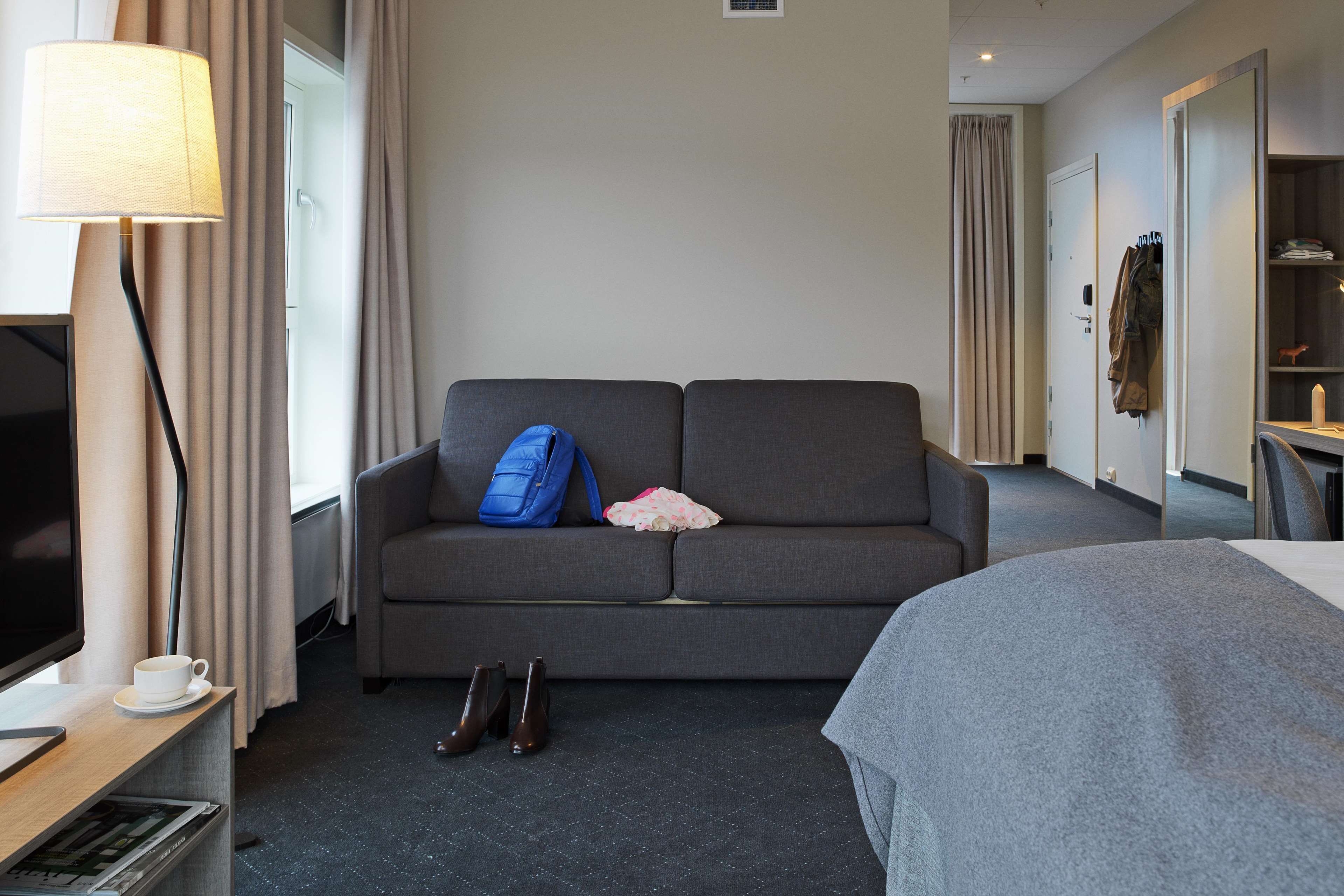 A hotel room with a grey sofa, a television, a lamp, and a bed.
