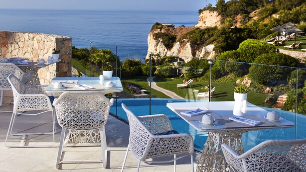 A poolside area with white wicker furniture, a clear view of the sea, and a landscaped garden.