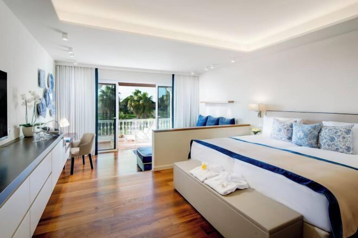 Een moderne hotelkamer met een groot bed, een zithoek, een bureau en een balkon met uitzicht op palmbomen.