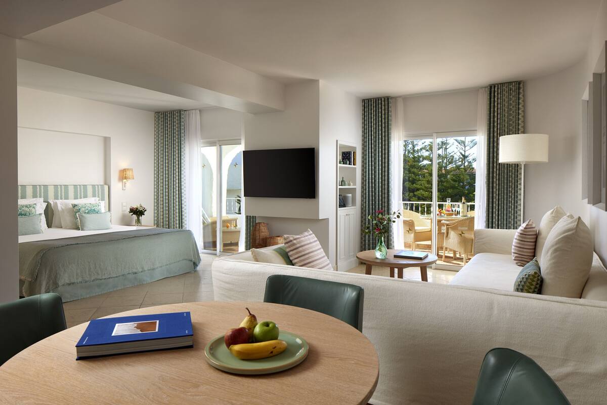 Een moderne hotelkamer met een bed, een slaapbank, een eettafel met fruit, een televisie en uitzicht op het groen.