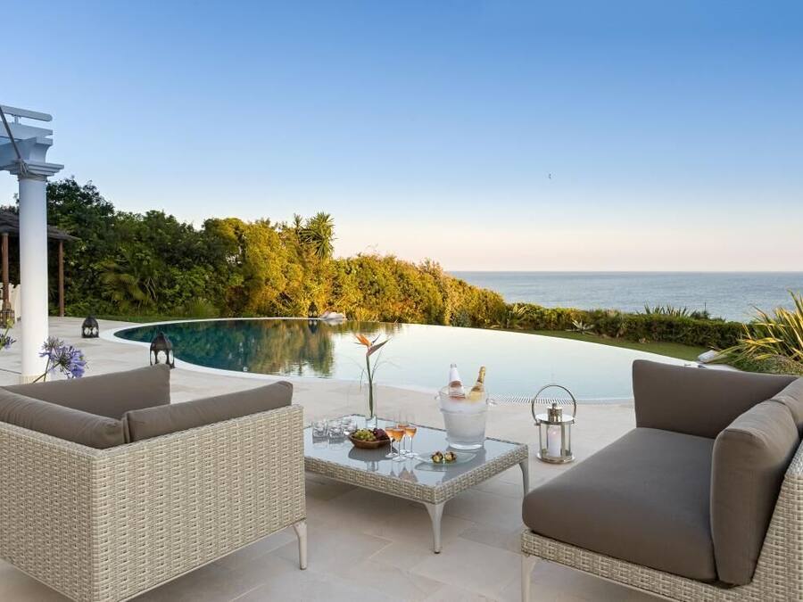 A poolside area with wicker furniture, a glass table, and a view of the sea.