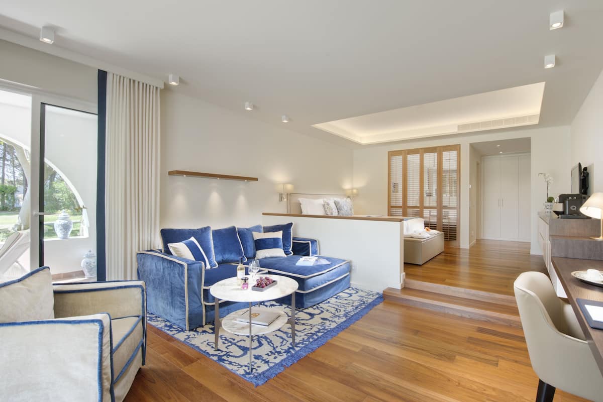 Een moderne hotelkamer met een blauwe bank, een ronde salontafel en een houten vloer. Er is een bureau met een lamp en een stoel.