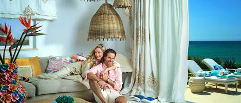 A couple seated on a sofa with a view of the ocean, a wicker chandelier, and a beachside lounge area.