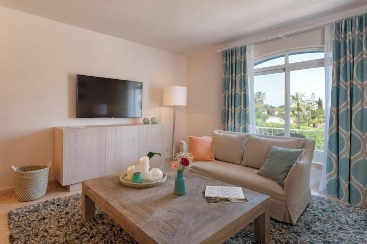 Een woonkamer met een bank, een bijzettafel met kaarsen en een vaas, een aan de muur bevestigde televisie, en een raam met gordijnen.