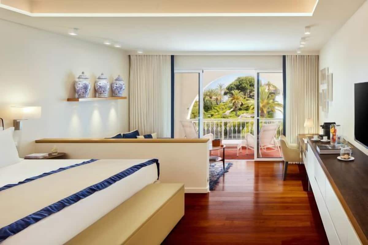 Een moderne hotelkamer met een groot bed, een zithoek, een balkon met uitzicht op groen en een televisie aan de muur.
